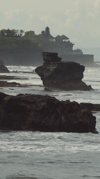 Soft Evening Tanah Lot Temple Silhouette On Cliff, Golden Haze And Calm Shallow Pools, Gentle Tide Lapping Basalt Shelf, Low Light Reflections And Meditative Travel Mood, Cultural Landmark Evoking
