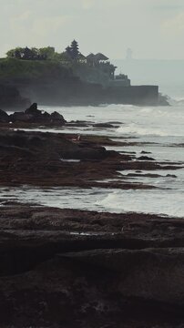 Stony Reef And Tanah Lot Temple Against Haze, Imagined Priest Conducting Quiet Ritual As Tide Sculpts Layered Rock, Steady Swell And Misty Horizon, Timeless Cultural Atmosphere With Muted Cinematic