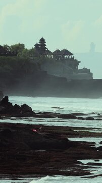 Stony Reef And Tanah Lot Temple Against Haze, Imagined Priest Conducting Quiet Ritual As Tide Sculpts Layered Rock, Steady Swell And Misty Horizon, Timeless Cultural Atmosphere With Muted Cinematic