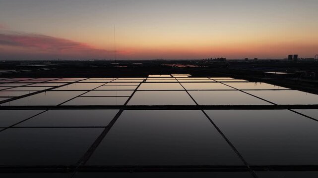Sunset over orderly salt pans with reflective water surfaces and distant city silhouettes
