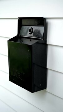 4K vertical video of a woman's hand opening a modern black wall-mounted mailbox attached to white house siding. The clean residential exterior and contemporary mailbox emphasize everyday homeownership