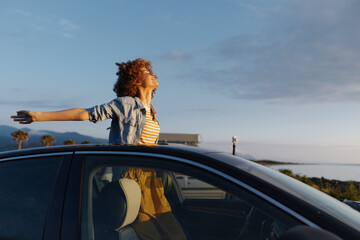 Obraz premium A woman leans out of a car window with arms outstretched, enjoying a sunny coastal drive. A carefree lifestyle moment capturing freedom, travel, and outdoor joy.