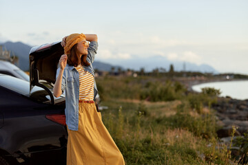 A woman lifestyle travel coast outdoors fashion stands beside an open car trunk on a sunny coastal road, enjoying a moment of freedom and light. © SHOTPRIME STUDIO