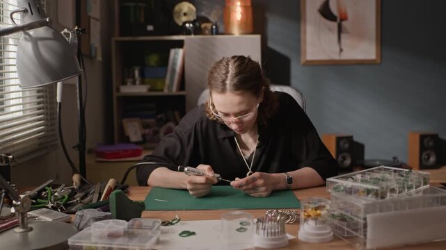 Slow motion shot of young Caucasian woman carving wax jewelry mold with rotary tool at workshop desk. Footage for promoting handmade jewelry brands, craft courses, or small business entrepreneurship