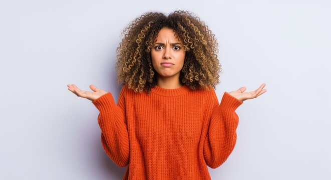 Young woman with curly hair in orange sweater shrugs with confused expression, palms open in questioning gesture against gray background. Perfect for expressing doubt or uncertainty.