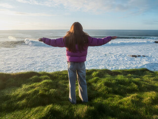Woman stands on grassy cliff near ocean waves. Red jacket contrasts with blue sky and sea. She smiles gently, facing horizon with calm. Sunlight highlights her hair and silhouette. Hands up in the air © mark_gusev