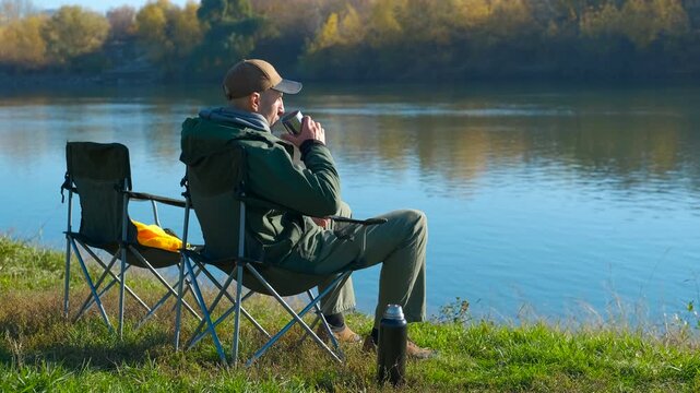 Fisherman relaxing and drinking hot tea by the river. Lone fisherman sitting in a folding camping chair on the riverbank, peacefully drinking a hot beverage from a thermos mug and enjoying the autumn