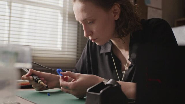 Close up of young adult Caucasian woman using soldering tool to carve blue wax ring model at workshop desk. Footage suitable for promoting handmade jewelry brands and artisan craft courses