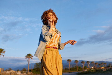 Fototapeta premium Young woman with curly hair stands outdoors in bright sunlight, wearing a striped top and mustard pants, smiling as she enjoys a carefree moment with palm trees in the background.