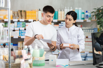 Fototapeta premium Young man choosing prescription medications with help of young female pharmacist in pharmacy