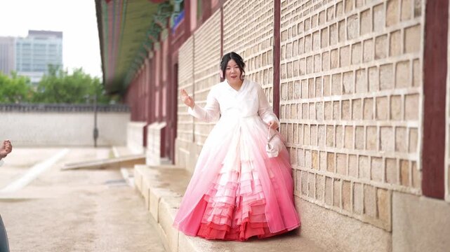 Young woman wears a traditional Korean gown. She leans against a brick wall, smiling gently. Her pink skirt flows with soft, layered ruffles. The scene is outdoors, near historic architecture