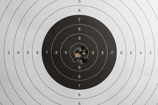 Paper target hanging on a bullet trap backstop wall in an indoor shooting range with an amazing sniper cluster of bullet holes right in the center ten score ring.