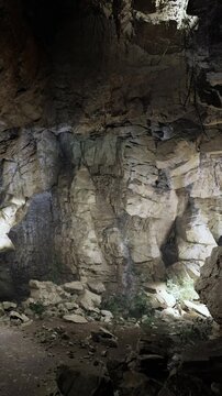 A group of people walks inside a rocky cave with light coming through the openings in the stone. The walls are rough and the ground is uneven. Plants grow in the rocks.