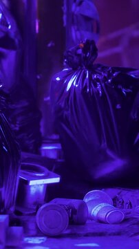 A collection of black trash bags is seen next to some discarded bottles in a dark urban location during nighttime when city lights create a purple hue.