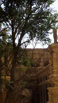 Visitors walk among old stone ruins during the day. A tall tree stands to one side, providing shade. The sun shines down on the historical site. People admire the old structures and carvings.
