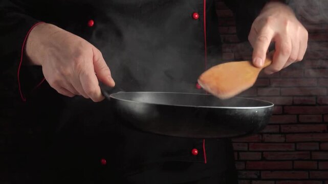 Chef in black uniform skillfully flips food in a frying pan, with steam rising and a brick wall backdrop enhancing the culinary atmosphere