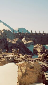 Visitors explore ancient stone structures in a vast desert. Sand covers the ground while rocky formations emerge. Bright sunlight illuminates the scene, revealing eroded details.