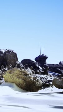 A building sits on rocky ground surrounded by snow. Two tall structures rise from the building against a clear blue sky. The landscape shows various large stones and frozen ground.