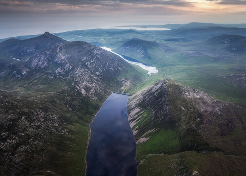 Aerial view of Ben Crom Reservoir nestled between rugged Mourne Mountains peaks with lush green slopes and calm water under a sunset sky in Newry, Northern Ireland, United Kingdom.