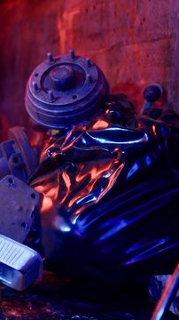 A shiny plastic bag rests on the ground surrounded by metal and mechanical parts in a dimly lit storage area. The blue and purple lights reflect off the surfaces creating a colorful scene.