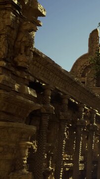 A stone structure shows intricate carvings on its walls and columns during daylight. The sun casts shadows, highlighting the design details of this historic site.