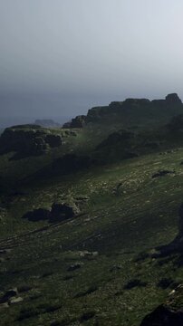 At a remote location, rocks and hills are illuminated by soft light. The terrain is uneven with patches of grass and distant cliffs can be seen. The atmosphere is quiet and vast.