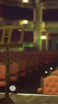 The theater stage shows a music stand in the front, with rows of empty red seats in the background. The lights create a warm atmosphere as the show is about to begin.
