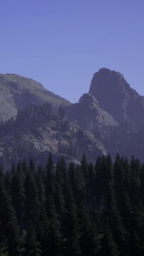 Tall mountains stand above a dense forest on a clear day. The peaks are rocky and steep. Green trees cover the area below, providing contrast to the sky. Nature is on display.