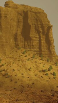 A tall rock formation rises from the ground in a dry area. Sparse vegetation surrounds the base. The sky has clouds present. The scene captures a remote natural setting.