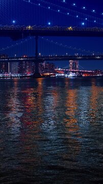 Dark riverscape shimmers with city lights at night. Low angle view at the Manhattan Bridge and the Brooklyn Bridge in New York City. Vertical video.