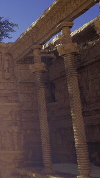 Visitors explore an ancient site with stone columns and intricate carvings. The sun shines on the weathered structures. Dust rises as people walk on the ground among the ruins.