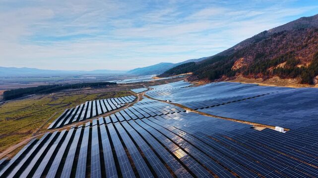 Solar farm along mountain slope and valley. Photovoltaic rows extend beside woodland and agricultural fields.