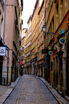 Lyon, France - 10.03.2026. A low-angle perspective looking down a charming, empty cobblestone street flanked by colorful multi-story Renaissance-era residential buildings, boutique shop signs, and tra