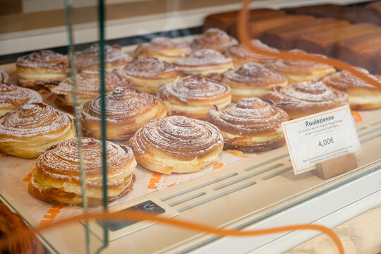 Rows of golden Roul&eacute;zienne pastries dusted with powdered sugar inside a glass display case at a French bakery. This close-up shot captures the spiral, laminated flaky viennoiserie rolls&mdash;made with butt