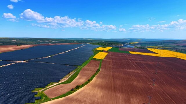 Solar arrays beside patchwork farmland and power lines, dark panels, tilled soil and distant hills below.