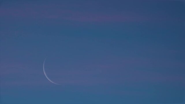 A slim crescent moon rises in a darkening sky past thin clouds tinted with the subtle pink of sunset.