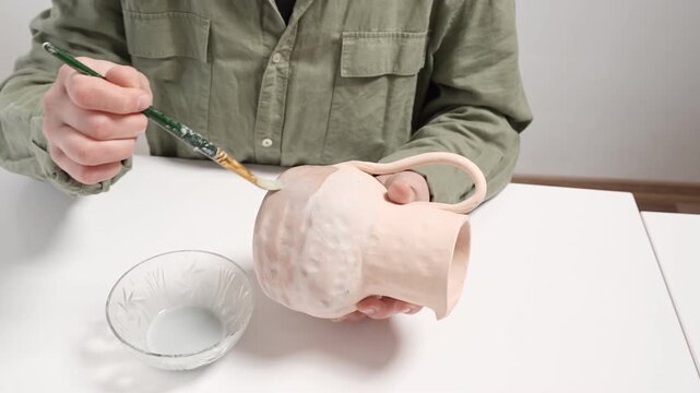 4k video showing an artisan glazing a bisque ceramic bowl with a wide studio paintbrush. Artist carefully applies thick glaze to the handmade clay vessel for a unique artistic finish