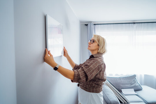 Side view middle aged woman putting painting picture in frame on the wall. Casual lady taking care of coziness in her new stylish apartment. Mature female decorating room. Moving and renovate house.