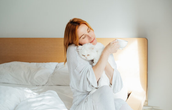 Smiling middle aged woman in pajama hugging cat pet and holding cup of coffee relaxing in bed in morning sunlight. Well being morning routine and daydreaming mood. Good morning habits.