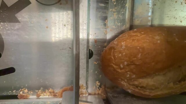 A top-down view shows a mechanical slicer cutting a loaf of artisan bread into uniform slices. The industrial setting features stainless steel surfaces with small crumbs scattered near the blades.