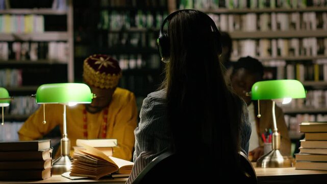 Back view of young student with headphones seated in moody library study room, desk with peers studying materials, reading and writing under green lamps with books, for education technology.