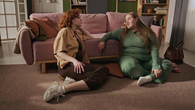 Caucasian young adult female friends, including plus-size woman, sitting on floor in living room, talking and laughing. Clip is suitable for body positivity campaigns and lifestyle advertisements