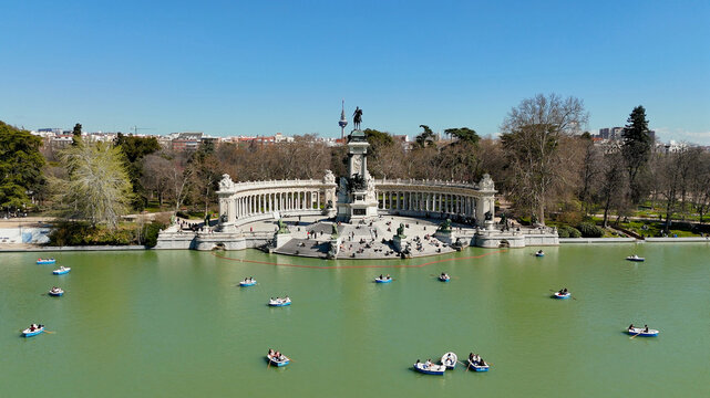 Wide drone view of the Monument to Alfonso XII overlooking the lake in El Retiro Park, Madrid, Spain, with rowing boats, calm green water, city skyline and large blue sky