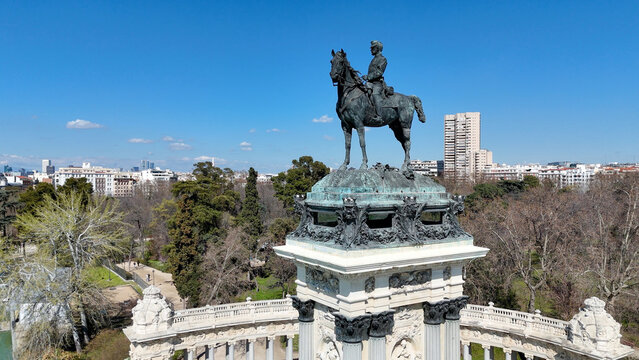 Drone photograph featuring the equestrian statue of King Alfonso XII atop the monument in El Retiro Park with Madrid skyline stretching across the background under a clear blue sky