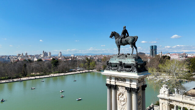 Wide aerial composition showing the Monument to Alfonso XII beside the boating lake in El Retiro Park with Madrid skyline visible in the distance on a bright sunny day