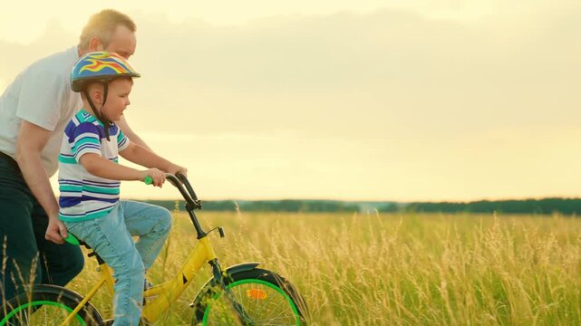 Dad helps child son ride bike. Father teaches child wearing safety helmet to ride bicycle in park. Child rides bicycle. Kid son dad play together, sun sky. Kid son dream learns to ride bicycle. Family