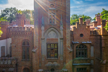 Fototapeta premium Abandoned Neo-Gothic Castle Facade Close-Up — Tudor Manor Pointed Arch Portal with Sun Flare, Decorative Spires and Crumbling Brick Walls | Historic Estate Detail