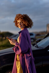 Confident woman in a purple coat stands beside a car, using a smartphone, showcasing casual lifestyle and chic fashion in warm sunset light, bright colors, relaxed pose and stylish mood