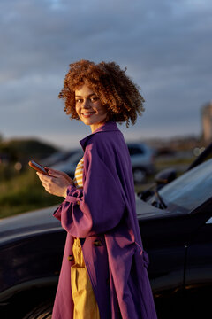 Confident woman in a purple coat stands beside a car, using a smartphone, showcasing casual lifestyle and chic fashion in warm sunset light, bright colors, relaxed pose and stylish mood