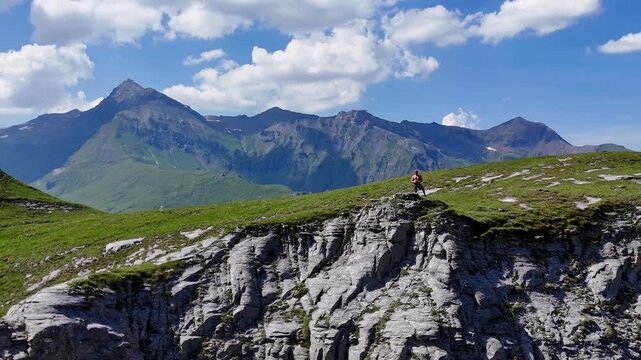 A spectacular aerial drone shot pulling back from a shirtless male hiker standing on a steep rocky cliff, flying away to reveal the immense scale of the green alpine ridges and majestic mountain peaks